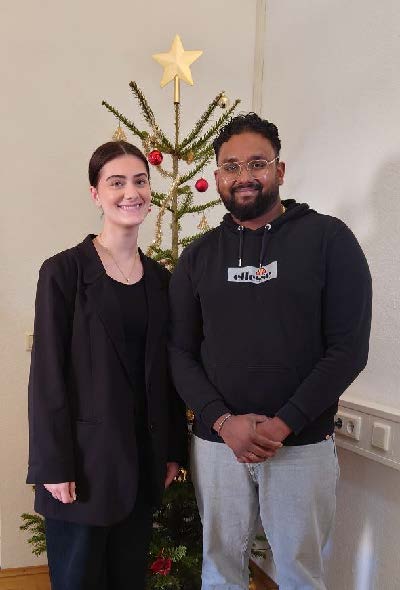 Bild von zwei jungen Menschen, die lächelnd vor einem einfachen geschmückten Weihnachtsbau stehen.